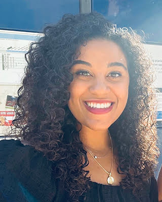 A person is smiling at the camera, with curly hair, wearing a dark top, and a necklace. Bright sunlight and building reflections are visible behind.