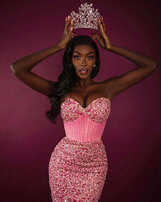 A person in a pink, jeweled dress poses against a burgundy background, gracefully holding a crown above their head with an elegant expression.
