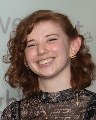 A person is smiling at the camera with curly auburn hair, wearing a black top with letter designs, and has multiple earrings. The background is neutral.