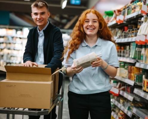 Two young people are stocking shelves in a grocery store, smiling and working together with one holding a box and the other reading a list.