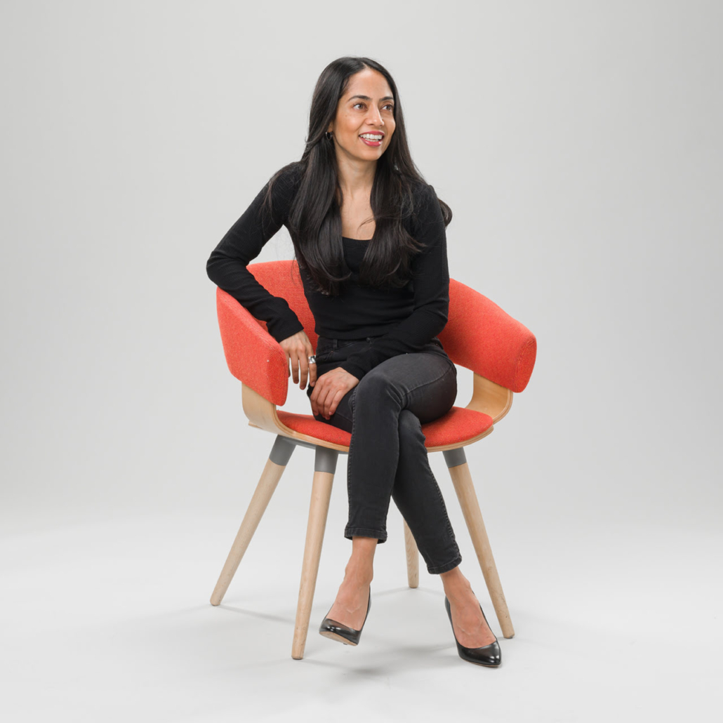 A person is sitting relaxed in a red and wooden chair against a grey background, wearing black attire and smiling slightly to one side.