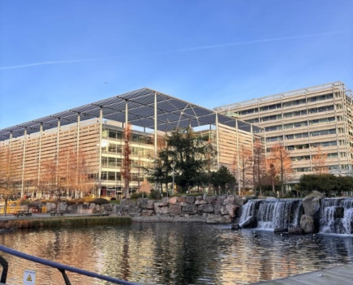 This image shows a modern urban landscape with cascading waterfalls, a reflective pond, contemporary buildings, clear skies, and a caution sign by the water.