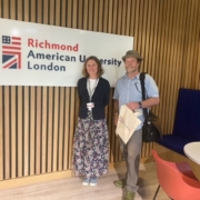 Two individuals are standing in a modern interior space with a sign reading "Richmond American University London" on a wooden slatted wall behind them.