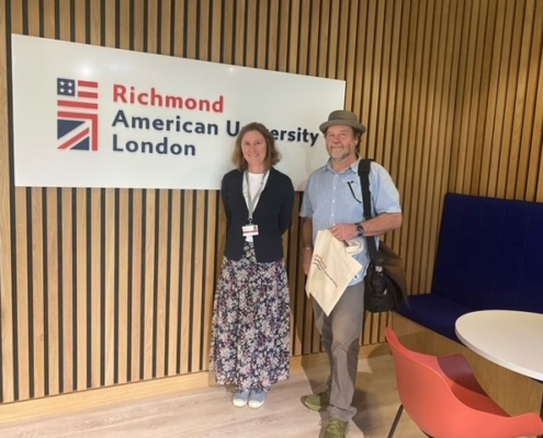 Two individuals are standing in a modern interior space with a sign reading "Richmond American University London" on a wooden slatted wall behind them.