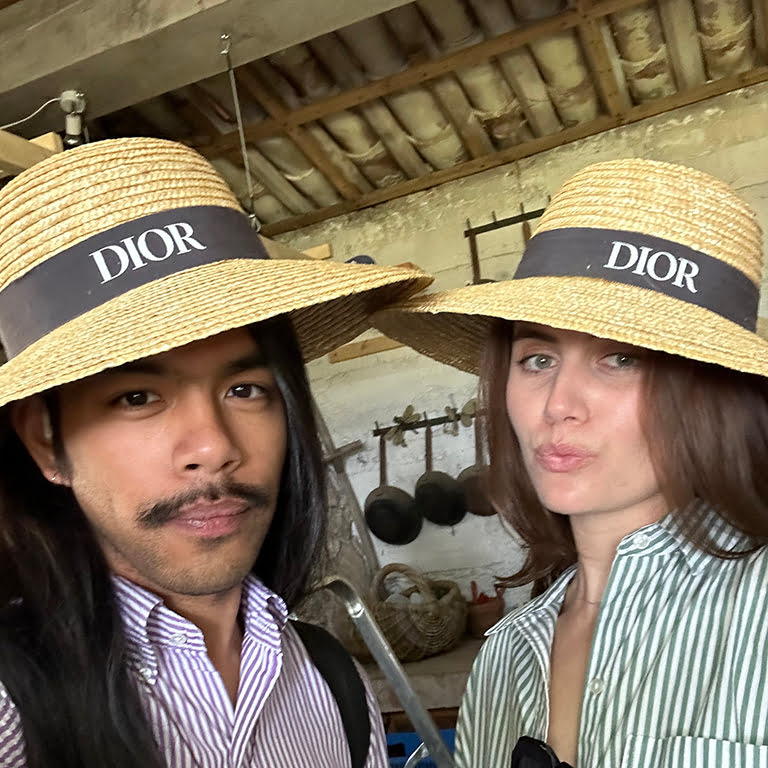 Two people wearing matching straw hats with "DIOR" bands pose closely, set against a rustic background with visible traditional kitchenware.