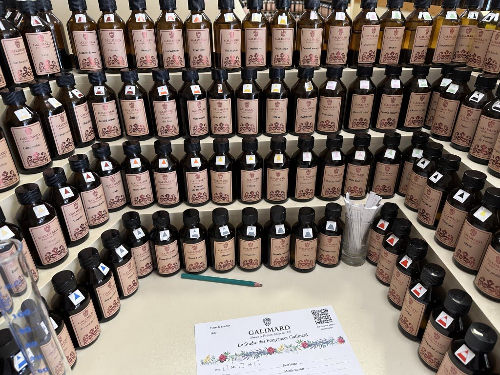 Rows of amber glass bottles labeled "GALIMARD" displayed on a table, alongside a paper with floral graphics and a pen, possibly in a perfume workshop.