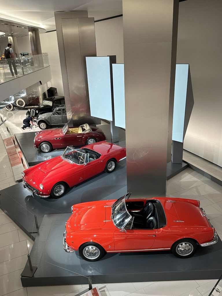 The image shows a car showroom with three vintage red cars on display, modern architecture, and a person standing in the background.