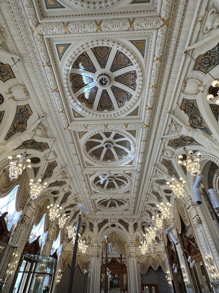 The image displays an ornate ceiling with intricate patterns, elegant chandeliers, and decorative windows, showcasing sophisticated architectural design and luxurious interior decor.