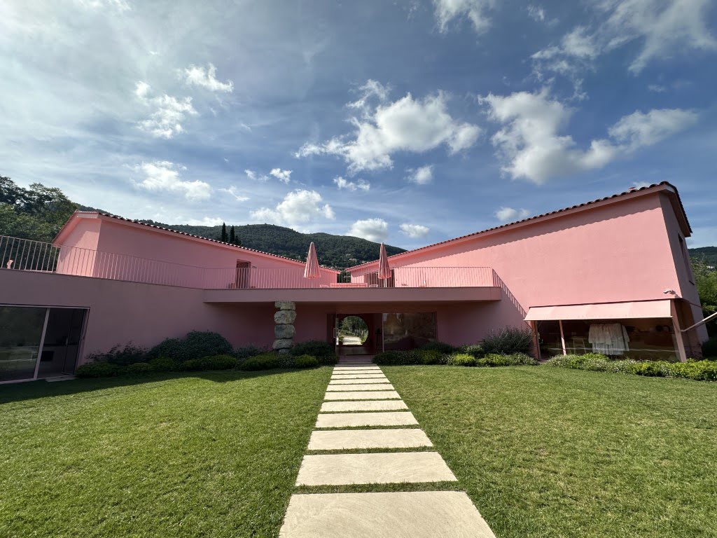 A unique pink building with a modern design, featuring a stepped path leading to it, surrounded by well-maintained green lawns under a cloudy blue sky.