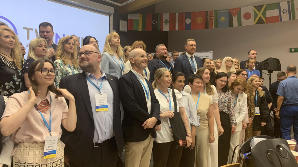 A diverse group of people pose for a photo in a conference room, wearing name tags, with international flags displayed in the background.