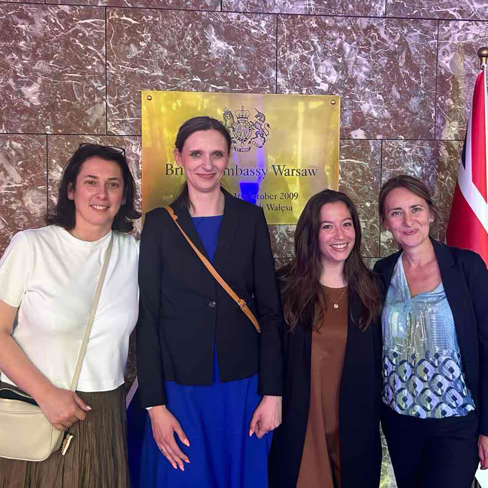 Four people are smiling for a photo in front of a British Embassy Warsaw plaque. They are dressed in business casual attire, with a marble background.