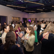 A group of people is networking at an indoor event, conversing and holding drinks, under ambient lighting with a digital screen visible in the background.
