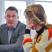 Two people in an indoor setting are engaged in a conversation. One person is attentively listening while the other speaks with a concerned expression.