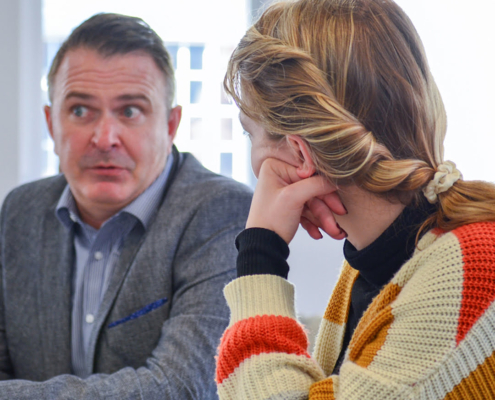 Two people in an indoor setting are engaged in a conversation. One person is attentively listening while the other speaks with a concerned expression.