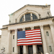 This image shows a large American flag hanging on the facade of a classical architecture building with sculptures and an arched pediment.