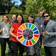 A group of seven smiling people posing outdoors, two holding a colorful circular object with a peace sign, in a sunny, green park setting.