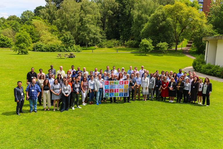 A group of people outdoors holding a banner with various colorful symbols, likely representing sustainability goals, gathered in a green park with trees.