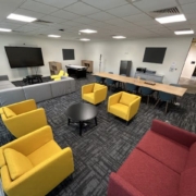 Modern office lounge area with colorful chairs, sofas, tables, multiple TV screens, and whiteboards, with carpet flooring and no people visible.