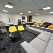 A modern office lounge with gray sofas, yellow and red chairs, a black coffee table, shelving units, and a desk with a computer.