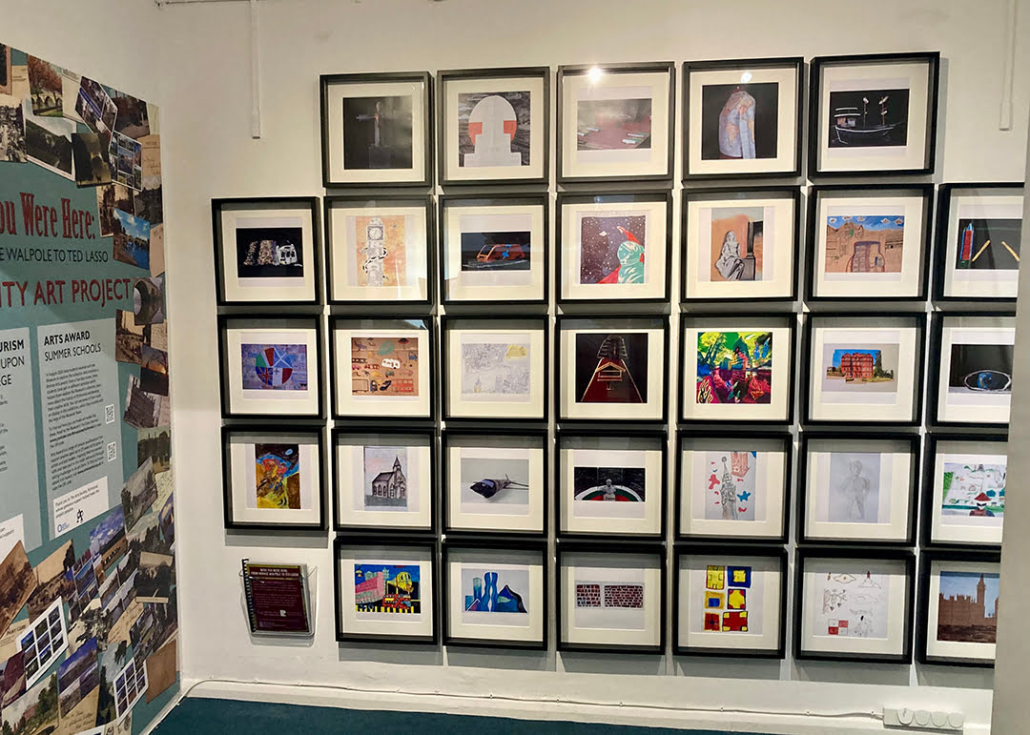 The image shows a wall with various framed artworks displayed in a grid pattern, alongside promotional material for a community art project.