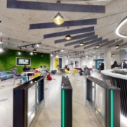 Modern office cafeteria with colorful furniture, a green living wall, and people working and socializing. Entrance security gates and contemporary lighting fixtures are visible.