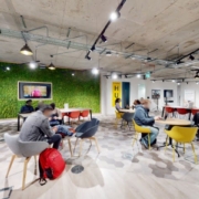 A modern, vibrant co-working space with people seated at various tables and couches, working on laptops, with a green accent wall and eclectic decor.