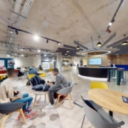 A modern, brightly lit interior with people seated at tables, an industrial ceiling, colorful furniture, and a central bar-like counter area.