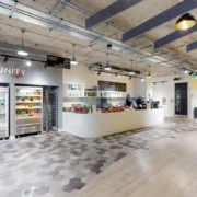 Modern office cafeteria with a contemporary design, featuring a snack bar, refrigerated display cases, minimalist decor, and exposed ductwork. A person is visible behind the counter.