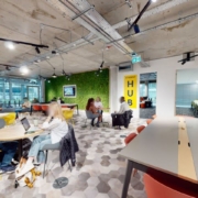 This image depicts a modern, brightly lit co-working space with various seating areas, people working at tables, a green wall, and exposed ceiling elements.
