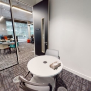 This is an office space with a person working at a desk. A small meeting area with a chair and table is in the foreground. Modern, bright interior.