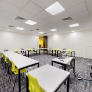 This image shows a bright, modern classroom with white tables, green chairs, and an interactive whiteboard system on the right. No people are present.