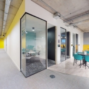 Modern office interior with yellow walls, glass-partitioned meeting rooms, exposed ceiling, colorful furniture, and a person sitting at a table.