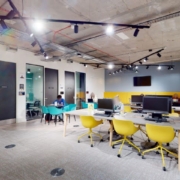 An open-plan office space with a modern design, featuring yellow chairs, wooden desks, exposed ceiling pipes, and people working on computers.