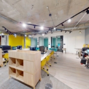 Modern office space with exposed ceiling, yellow accents, computer workstations, wooden bookshelves, and a group of people in a meeting.