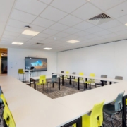Modern conference room featuring a long table with lime green chairs, white walls, a digital screen, blinds on the window, and carpeted flooring.