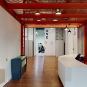 An industrial-style corridor with an exposed red ceiling grid, wooden floors, white walls, and a reception desk. There is one person sitting in the background.