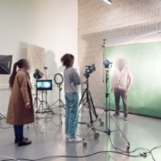 A photo studio with a green screen, lighting equipment, two people setting up, and one person standing in front of the screen, preparing for a shoot.