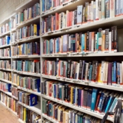 The image shows a library or bookstore aisle with tall, white shelves packed with an array of colorful books on wooden flooring, and no people.