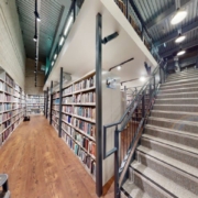 This image shows an interior of a library with bookshelves lined with books. A staircase in the center leads to an upper floor with more shelves.