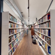 A person stands between rows of bookshelves in a library, browsing through books. Fluorescent lights illuminate the space with wooden flooring and white shelves.
