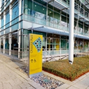 A modern glass facade building with metal framework, clear skies, and a yellow informational sign at the entrance, with people walking nearby.