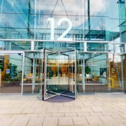 Modern building entrance with revolving doors, large glass façade, and the number 12 prominently displayed. Paved pathway leading to the entrance.
