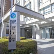 The image shows a signpost with Japanese characters in front of a modern building on a sunny day, with clear blue skies and manicured green bushes.