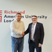 Two people are shaking hands in front of a wall with the "Richmond American University London" sign. They are smiling, wearing business attire.