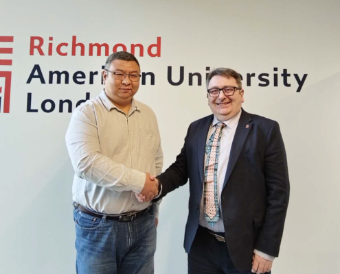 Two people are shaking hands in front of a wall with the "Richmond American University London" sign. They are smiling, wearing business attire.