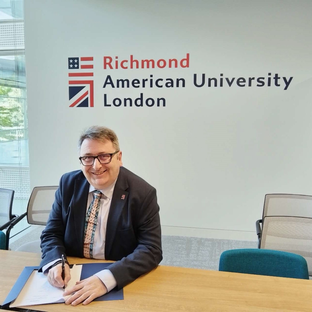 A smiling person is seated at a table, signing a document, in an office with "Richmond American University London" and flags on the wall behind.