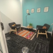 Small room with teal accent wall, two chairs, colorful rug, and framed artwork. A floor lamp and small table create a cozy atmosphere.