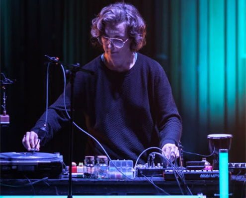 A person using DJ equipment, illuminated by teal and purple lighting, performs on a dimly lit stage with a microphone and headphones.