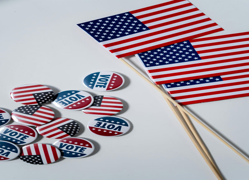 The image shows small American flags and vote buttons with patriotic designs arranged on a white surface, symbolizing a voting and election theme.