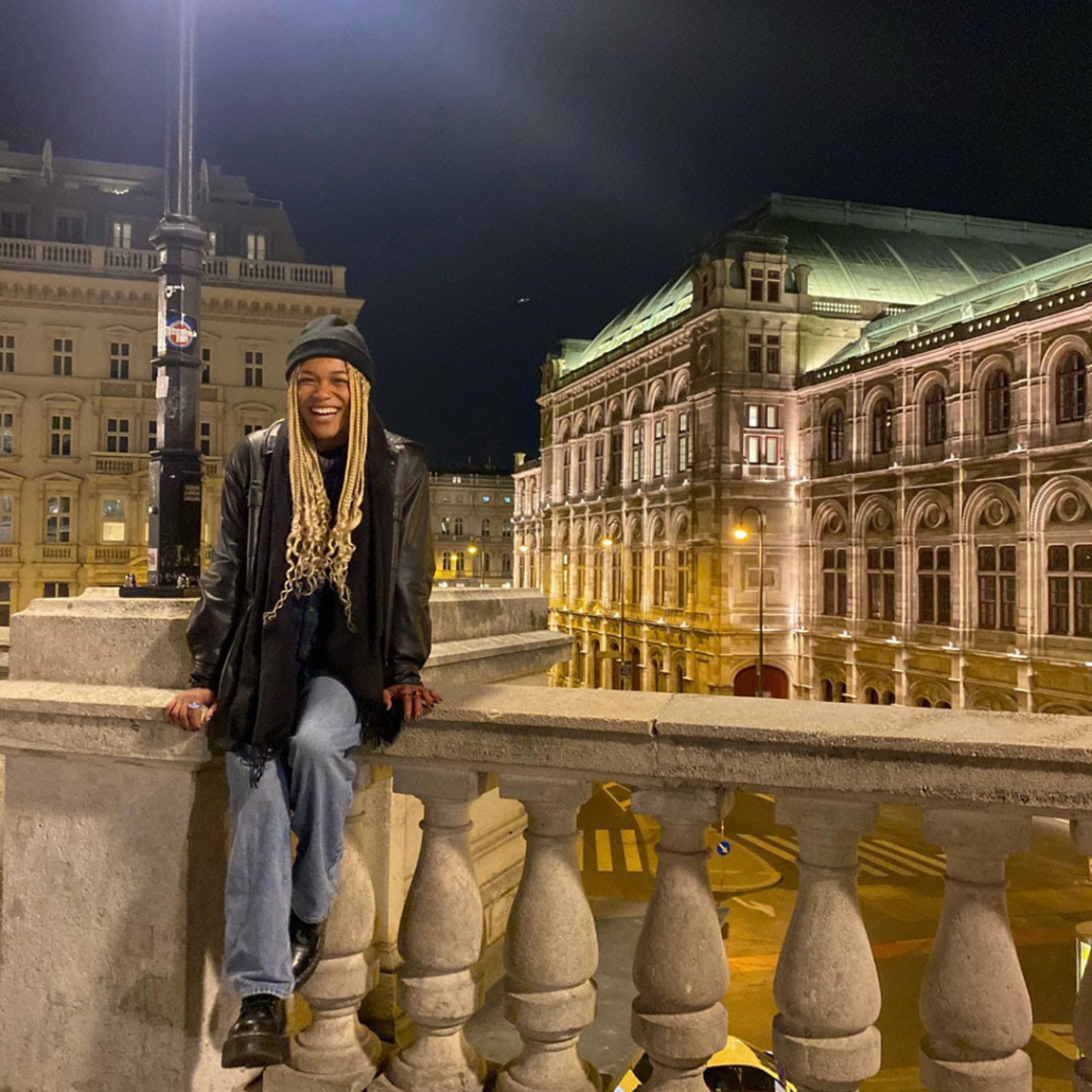 A person sits on a stone railing at night, smiling. The illuminated Vienna State Opera is visible in the background.